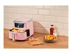 Pink air fryer with fries in the basket, a bowl of fries, ketchup, and salt on a wooden counter.