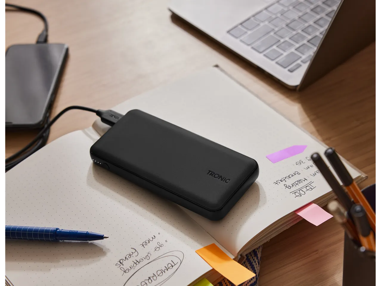 A TRONIC power bank charges a smartphone on a notebook next to a laptop and stationery.