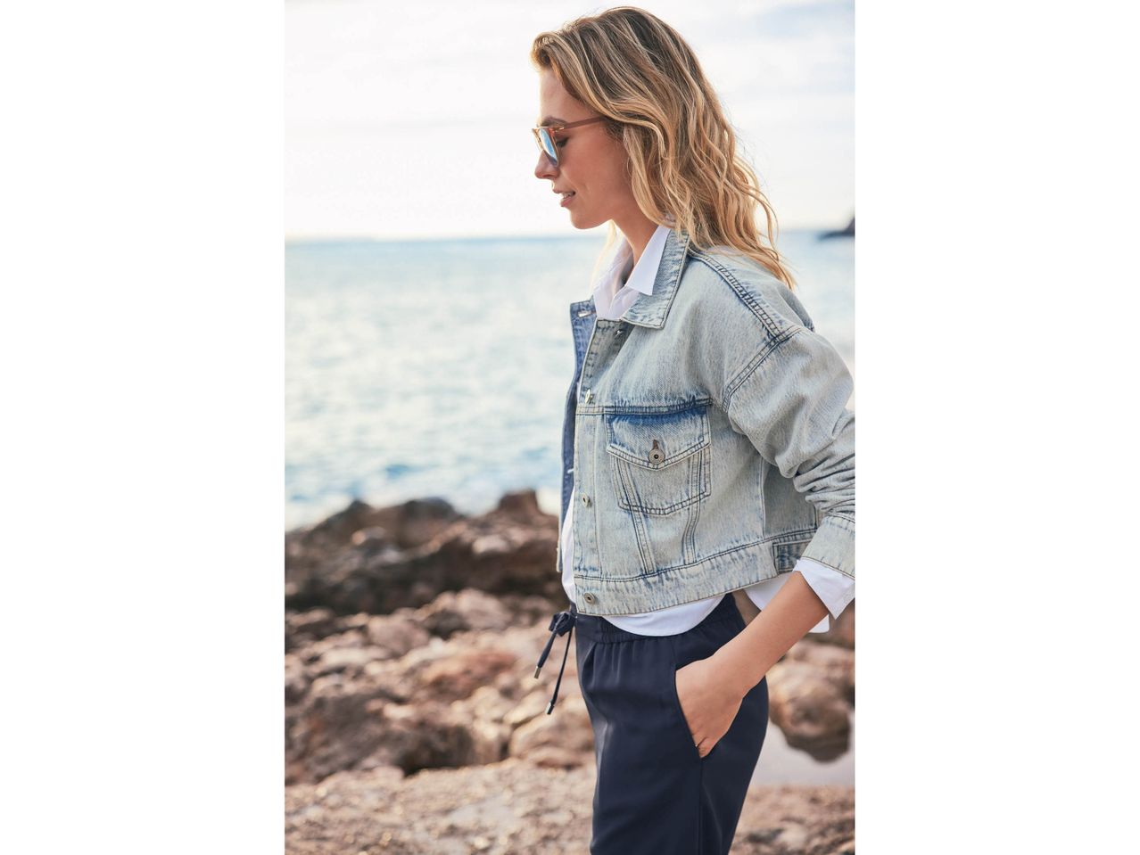 Donna in giacca di jeans, pantaloni blu e occhiali da sole sulla spiaggia.