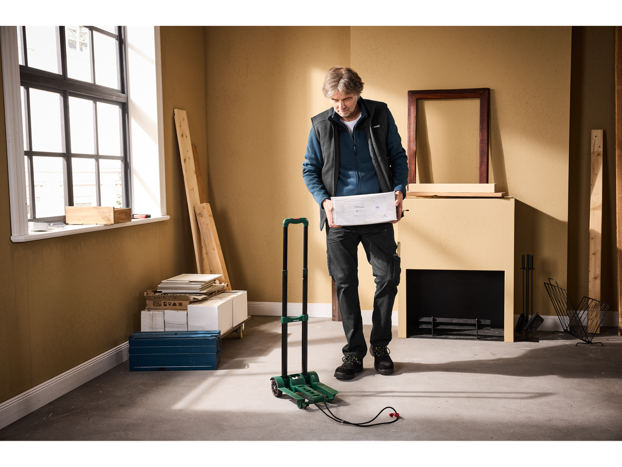 Man holding a box, standing next to a hand truck in a room under renovation.
