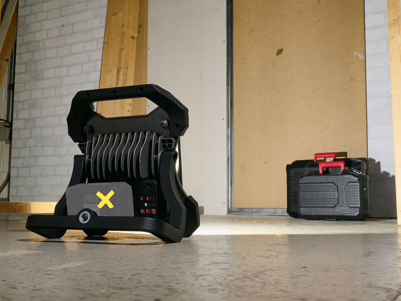 A Parkside work light illuminates a concrete floor in a workshop, near a tool case.