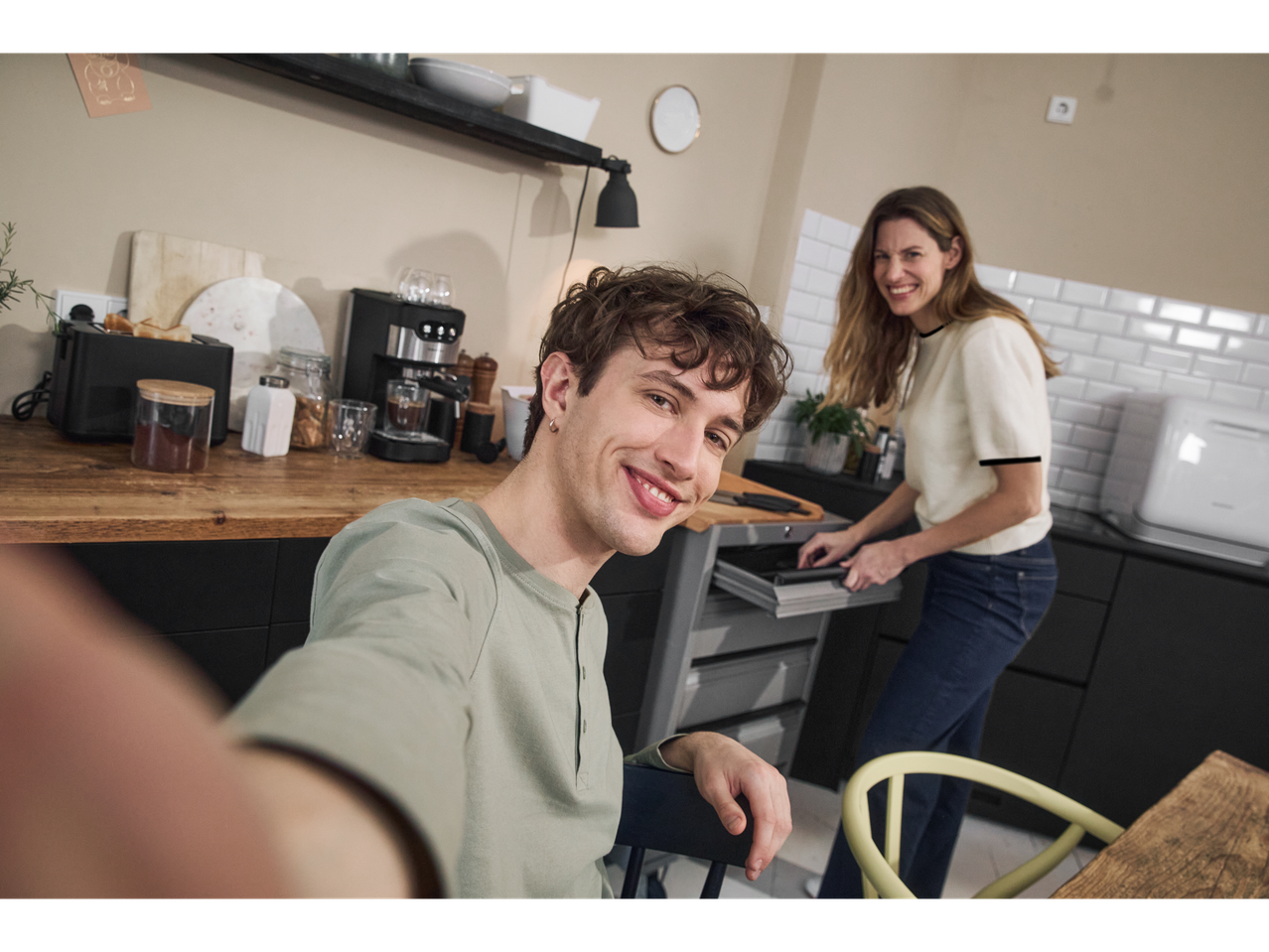 Young couple in a modern kitchen, taking a selfie with an espresso machine.