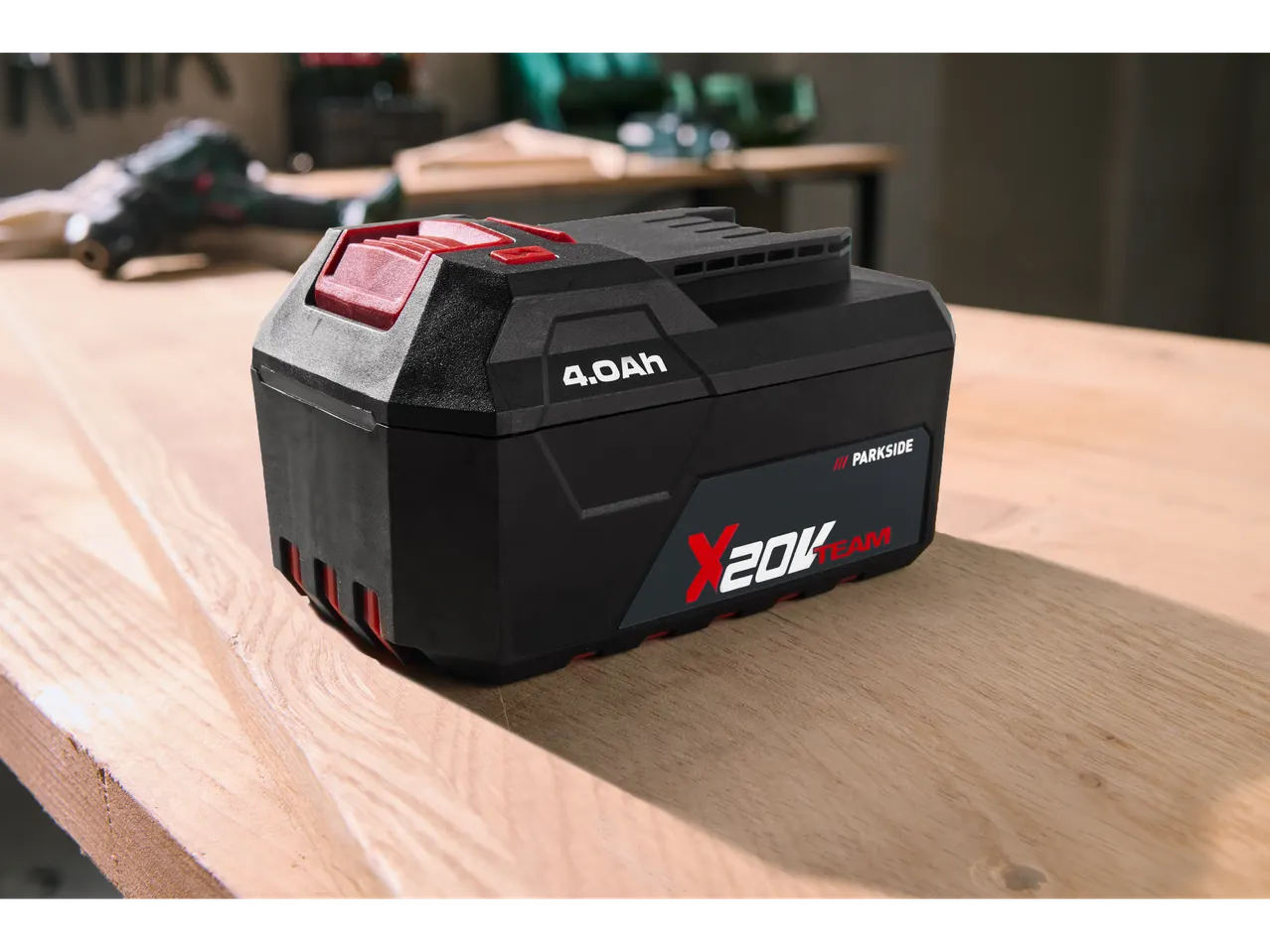 Батерия Parkside X 20 V Team 4.0 Ah на дървена маса, с размазани инструменти на заден план.