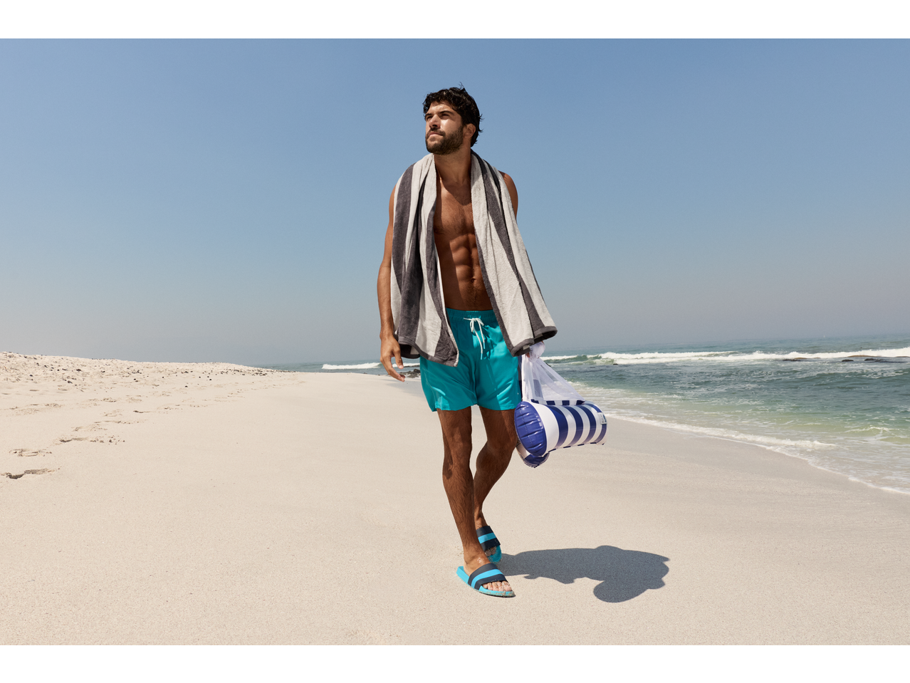 Homem com calções de banho CRIVIT e toalha na praia, a carregar um colchão insuflável.