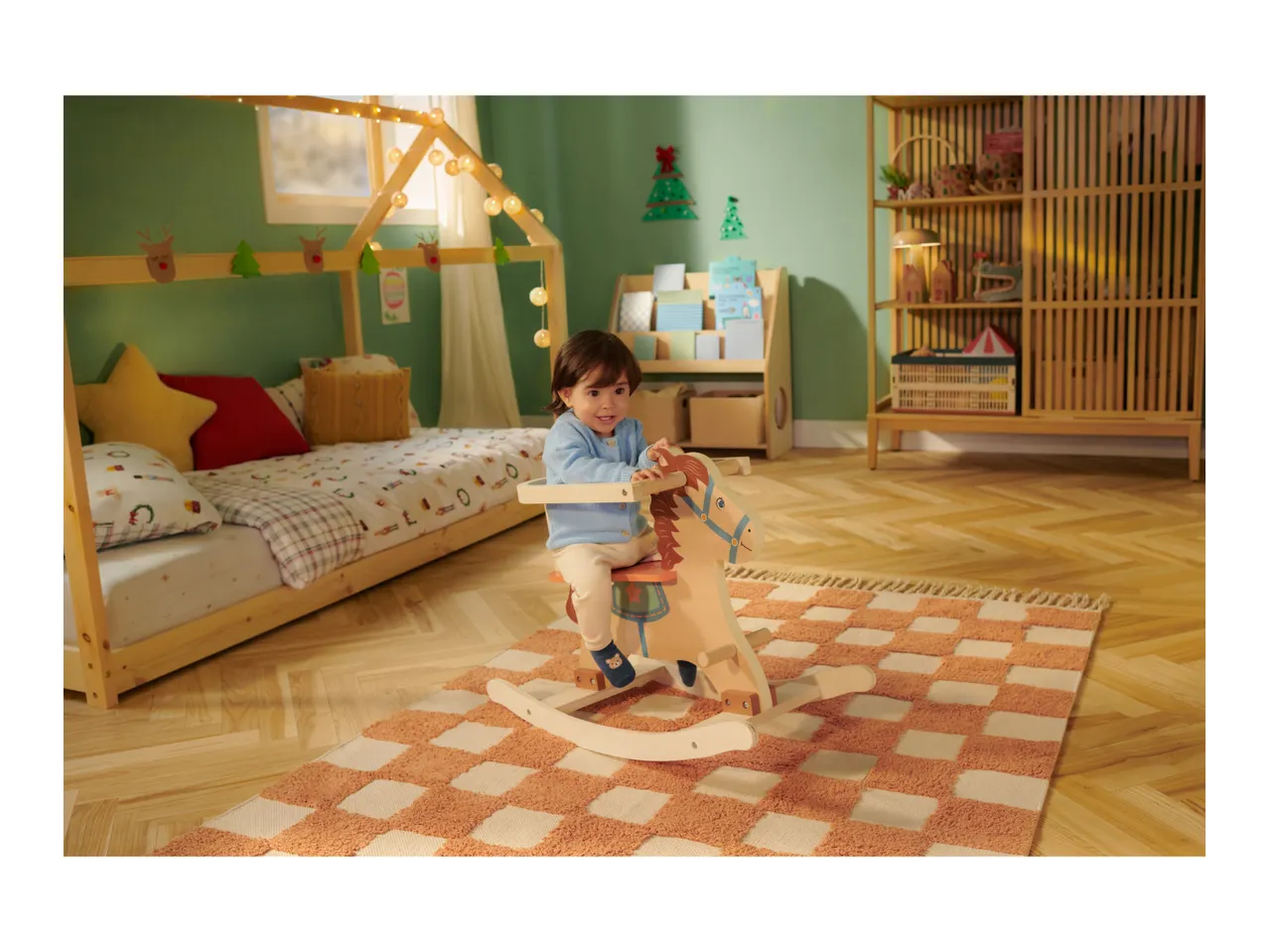 Toddler on a wooden rocking horse in a Christmas-themed bedroom with a house bed and rug.