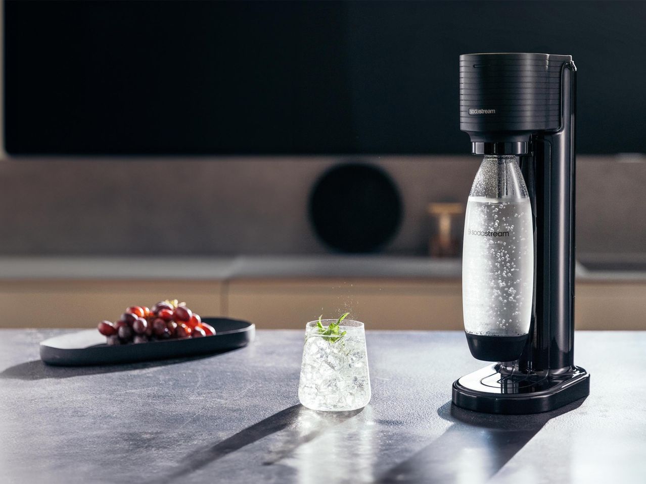 Fekete szódagép szénsavas vízzel, pohár jéggel és mentával, valamint tálca szőlővel egy konyhapulton.