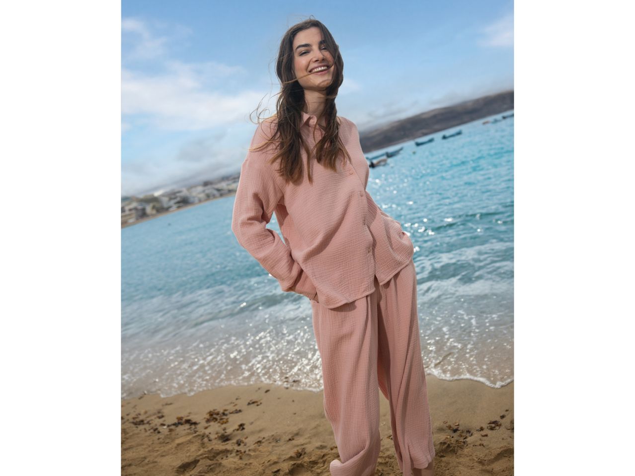 Woman in a pink muslin shirt and trousers set, smiling on a beach.