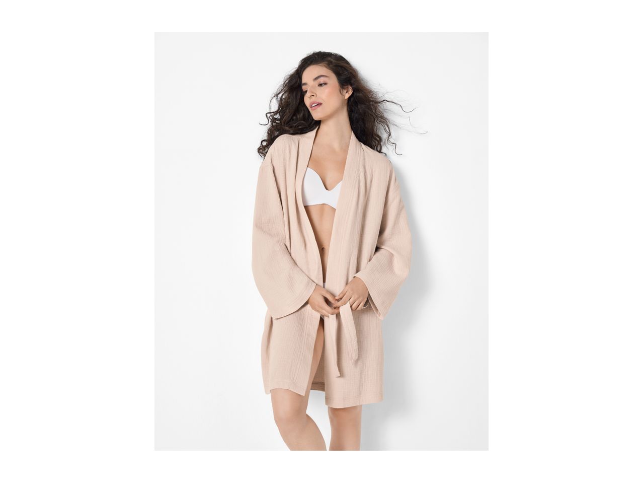 Woman in a light pink bathrobe and white bra, with long curly hair, looking to the side.