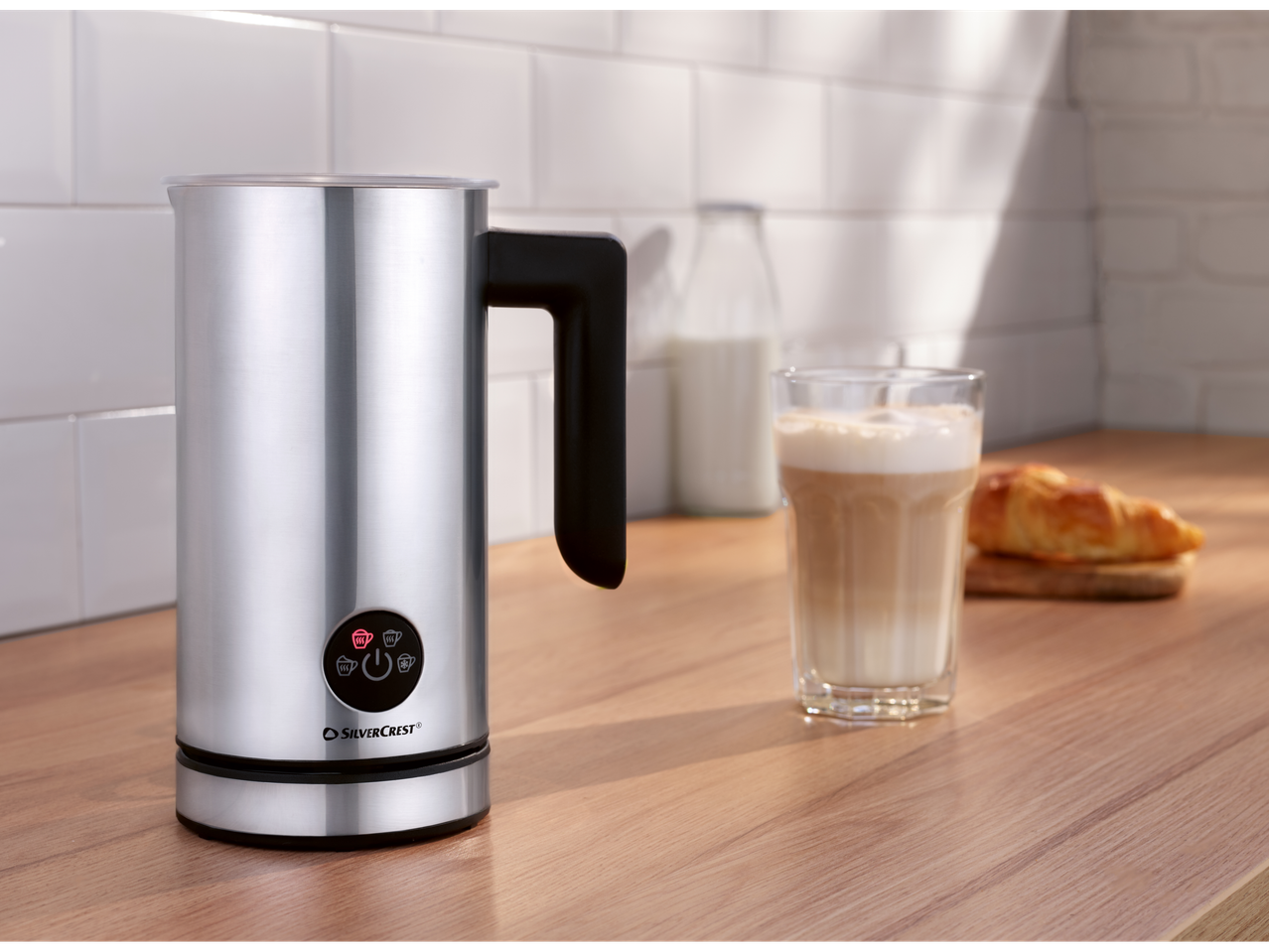 Silvercrest aparat za penušanje mleka na drvenoj radnoj ploči sa late kafom i kroasanom.