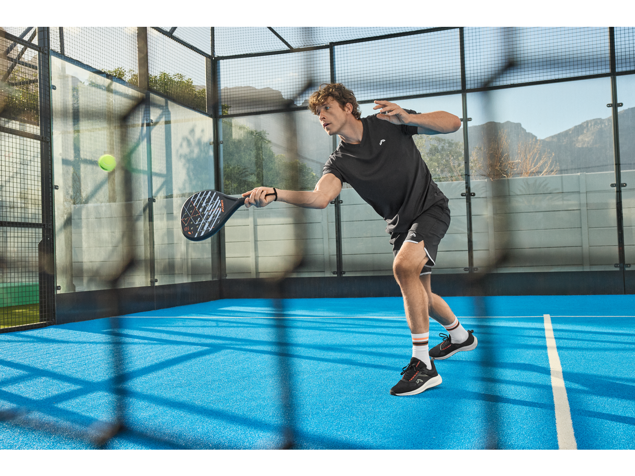 Man in zwarte sportkleding speelt padel op een blauw veld met een racket.