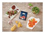 A blue packet of sun-dried tomatoes surrounded by steak, salad, bread, and garlic on a wooden table.