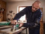 A man wearing safety glasses and earplugs uses a Parkside Paint Stripper to sand a wooden frame.