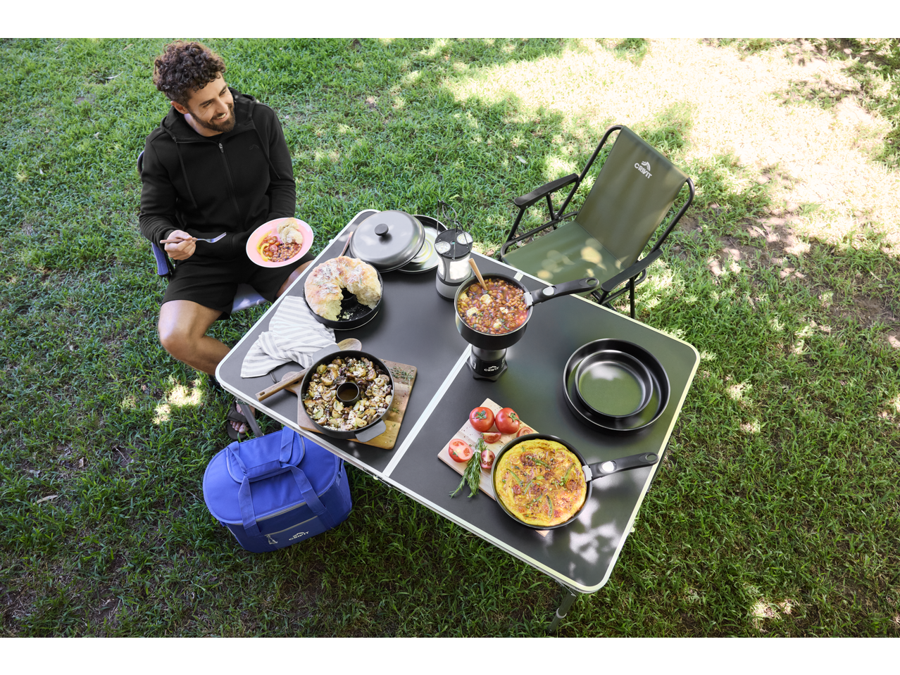 Overhead view of a man enjoying a camping meal with a portable table, chair, and various dishes.