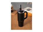 Black travel mug with a handle and straw on a wooden table, with a book and coffee maker in the background.