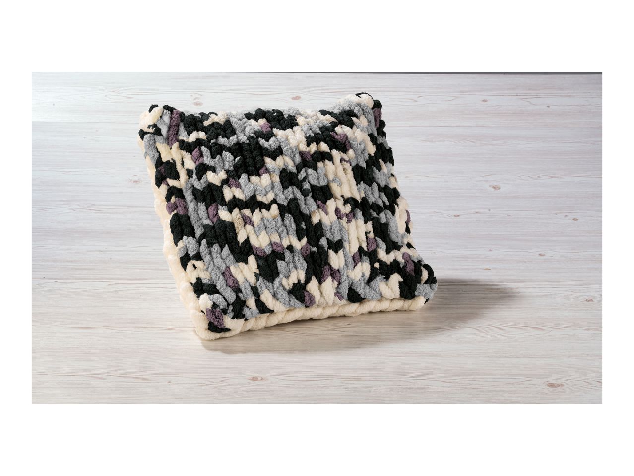 A chunky knit pillow in black, white, gray, and purple on a light wooden surface.