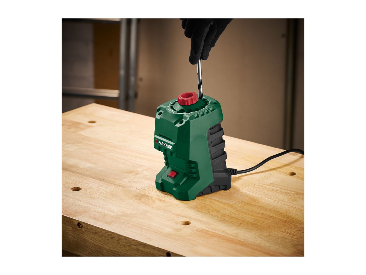 A person in black gloves sharpening a drill bit using a green and black drill sharpener on a wooden workbench.