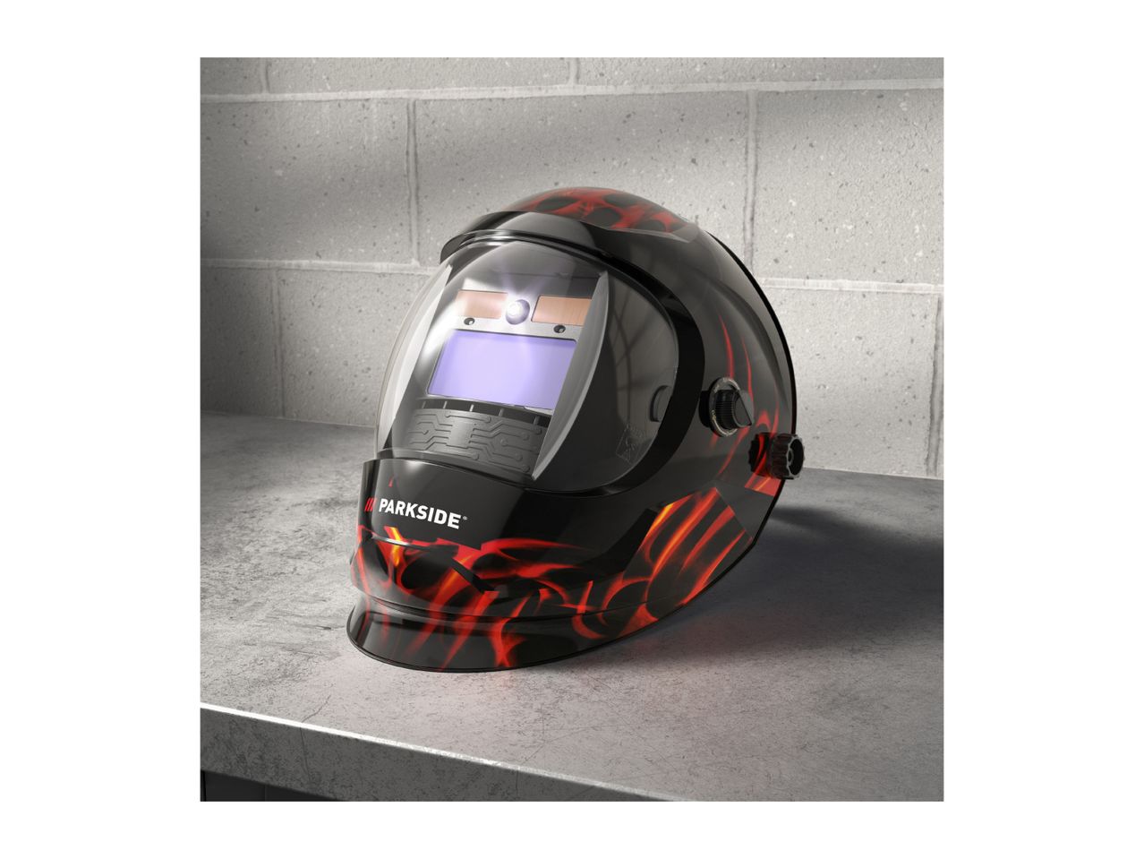 A Parkside welding helmet with a flame design sits on a concrete surface.