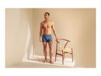 Man in blue boxer briefs standing next to a wooden chair in a sunlit room.