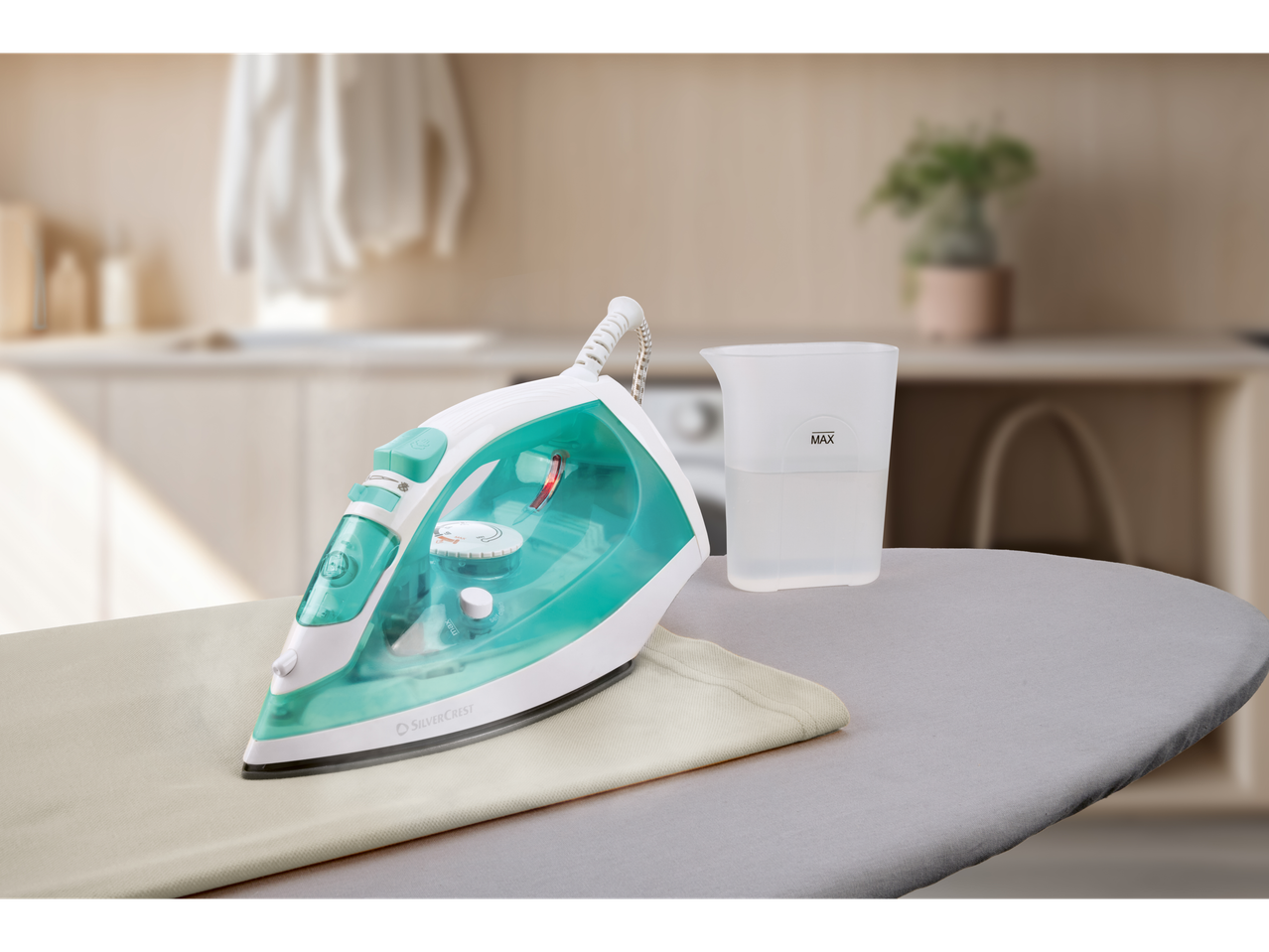 Silvercrest® parna pegla u tirkiznoj i beloj boji, postavljena na dasku za peglanje sa mernom čašom.