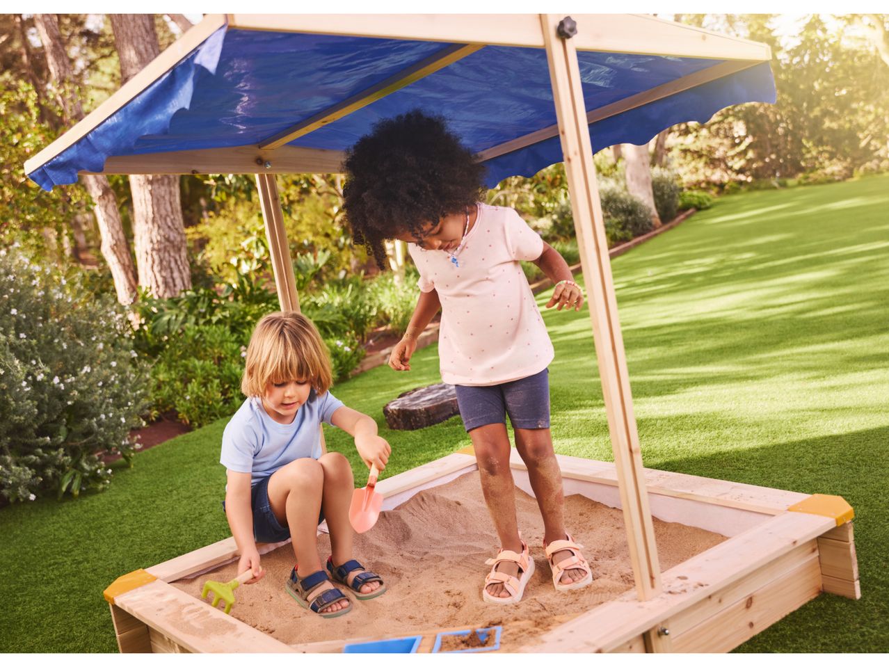 Two children playing in a wooden sandbox with a blue canopy in a garden.