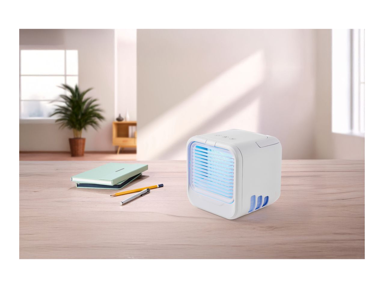 Portable air cooler on a wooden desk with notebooks and pens in a bright room.