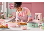 A baker decorates a pink cheesecake in a retro-style kitchen using pastel-colored baking tools.