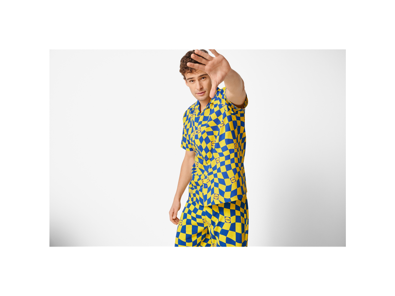Man in a blue and yellow checkered shirt and shorts, holding his hand up to the camera.