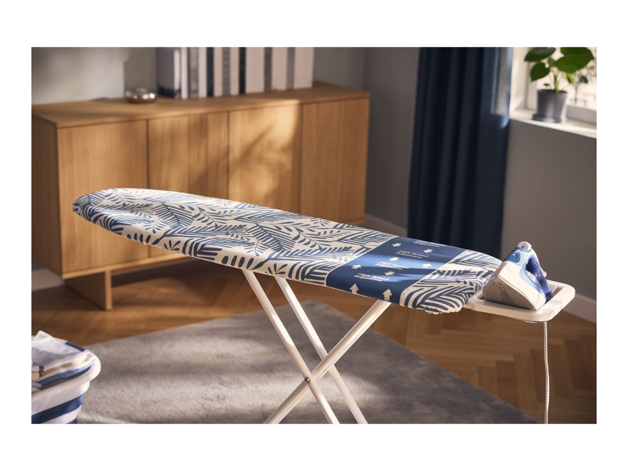 Ironing board with a blue and white leaf pattern, an iron, and a laundry basket in a living room.