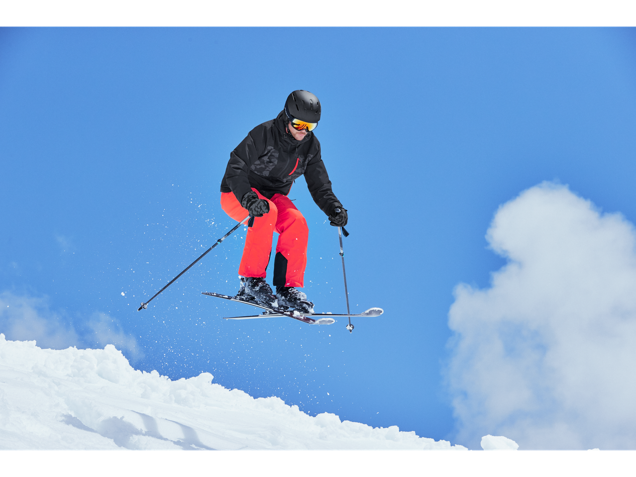 Suusataja CRIVIT jope ja punaste pükstega õhus hüppamas.