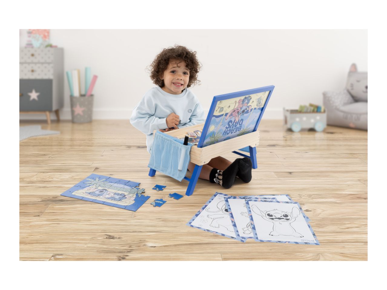 Happy child playing with a blue activity desk, puzzle, and coloring sheets on a wooden floor.