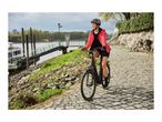 Woman in a red jacket and helmet riding an e-bike on a cobblestone path by a river.