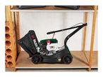 Petrol lawnmower with grass catcher bag on a wooden shelf in a storage area.