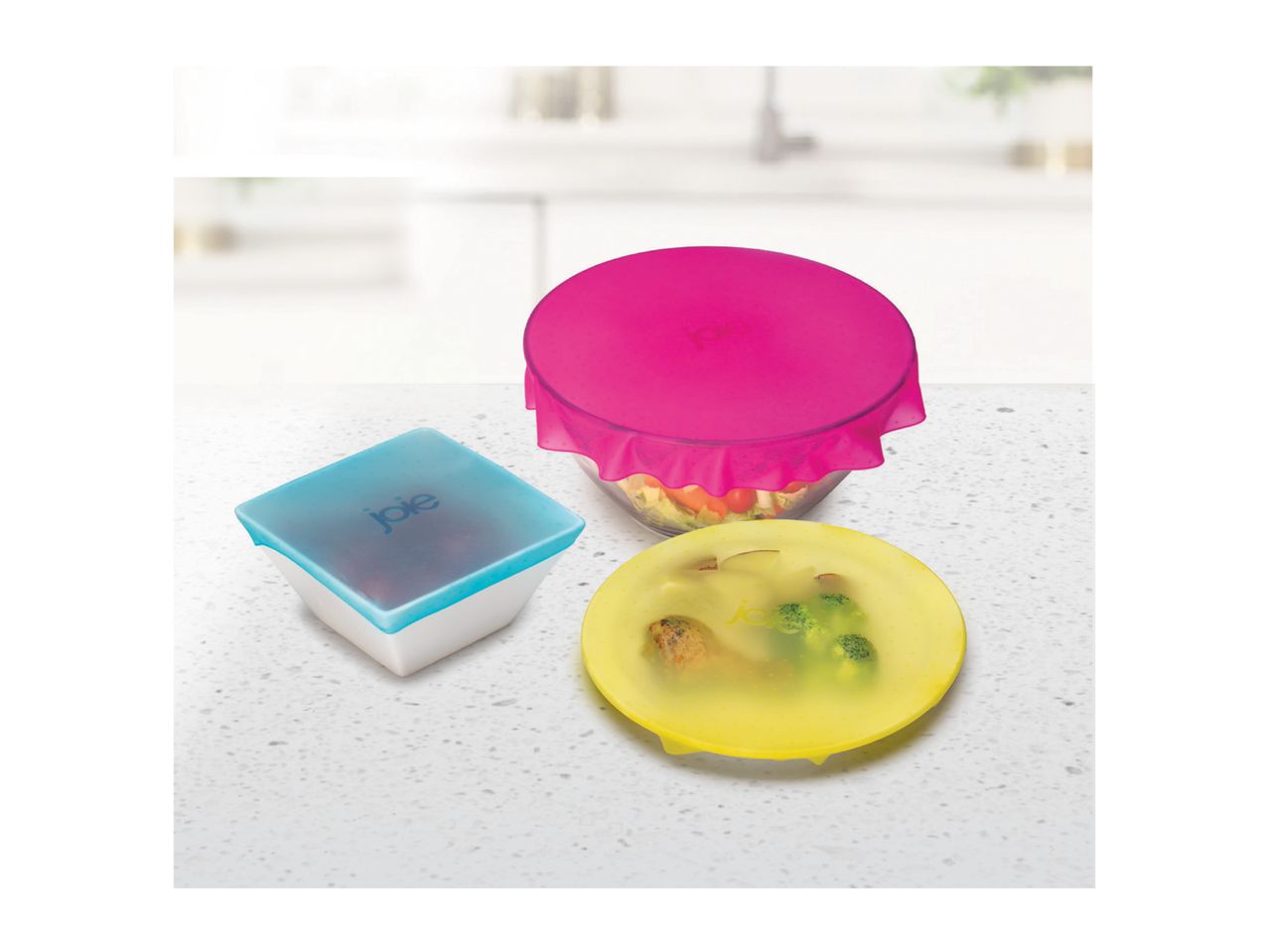 Three colorful silicone food covers in pink, blue, and yellow, covering bowls of food on a kitchen counter.