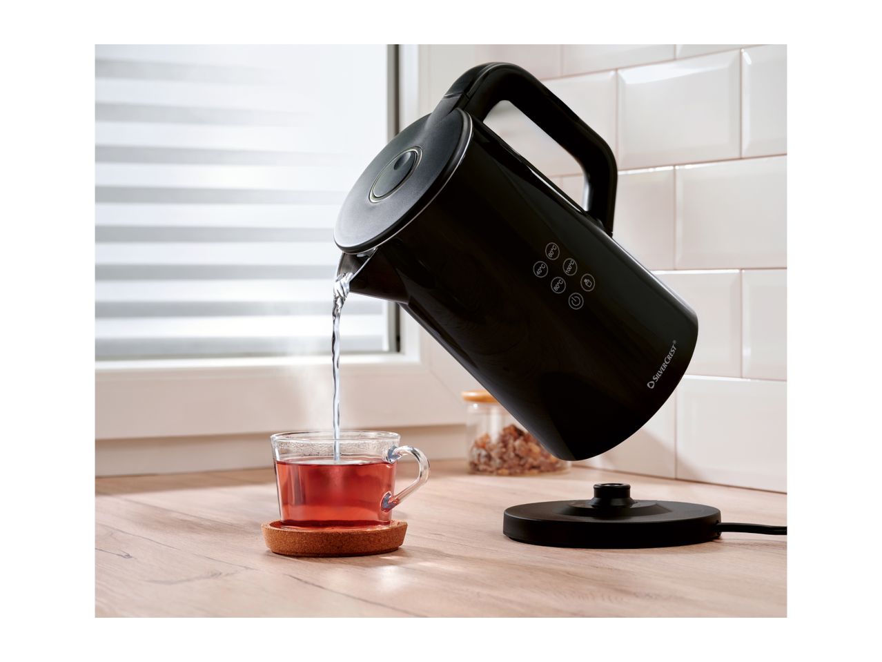 Black electric kettle pouring hot water into a glass cup of tea on a wooden counter.