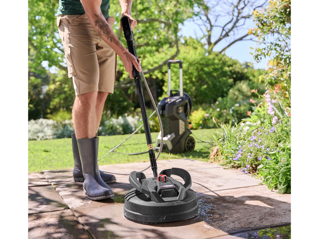 Man in rubber boots cleaning a patio with a pressure washer and surface cleaner.