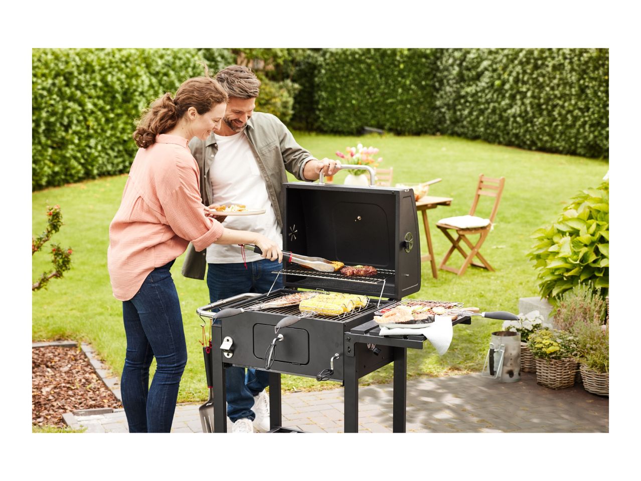 Couple grilling outdoors with a charcoal barbecue, cooking corn, fish, and burgers.