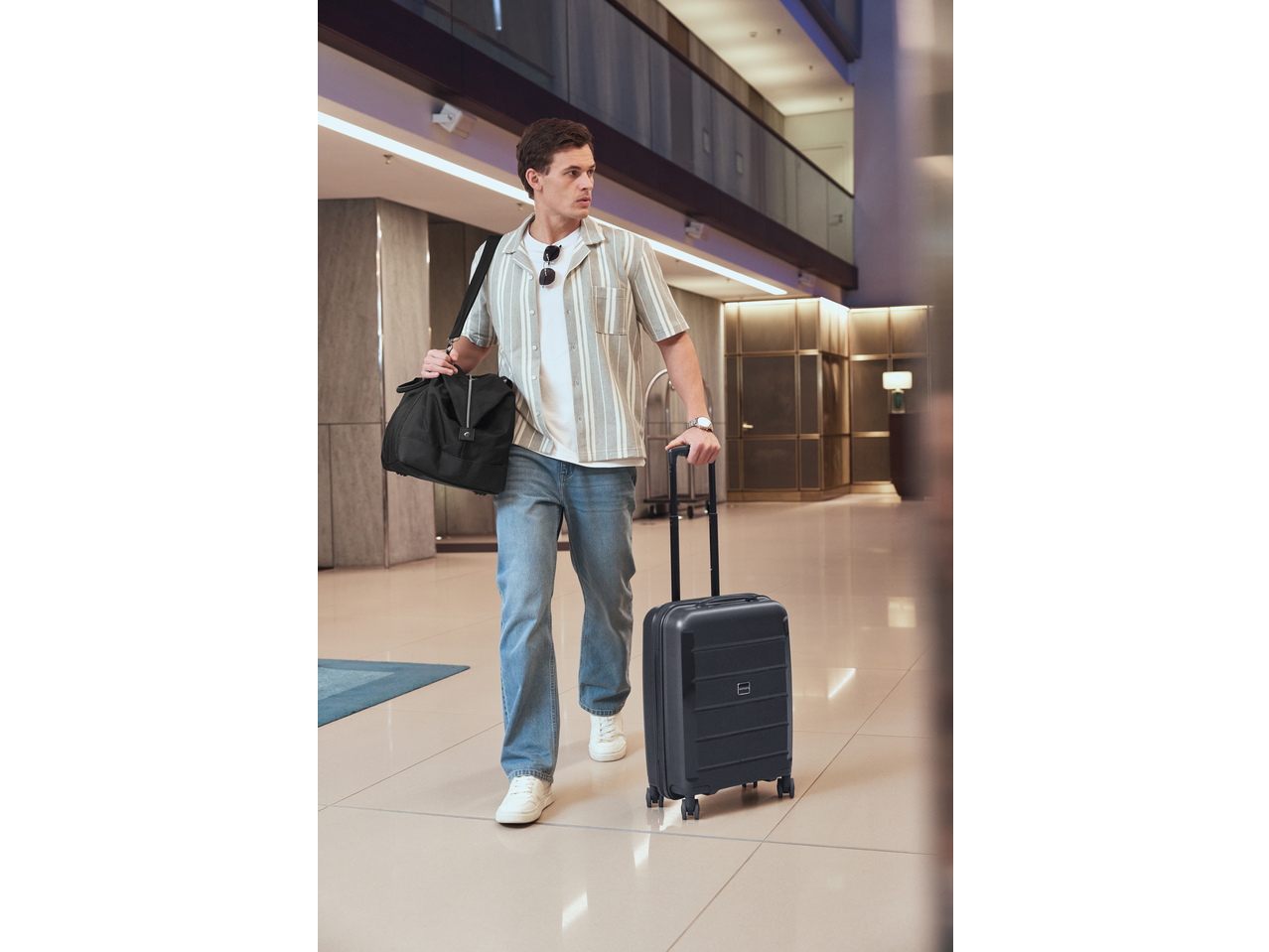 Homem de camisa às riscas e jeans, puxando uma mala ESMARA e carregando uma mala de viagem.