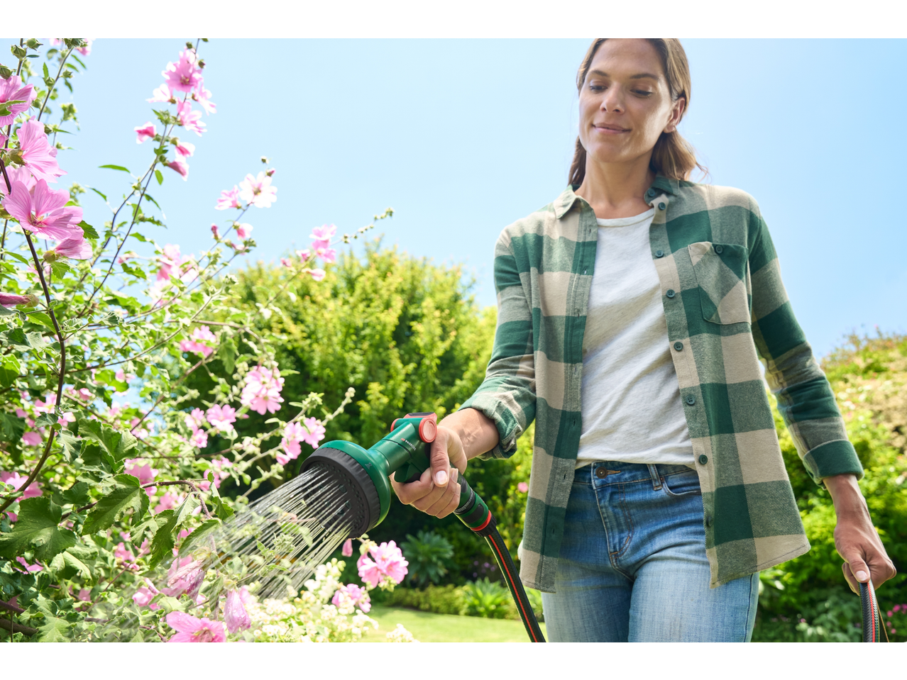 Woman watering pink flowers with a garden hose and spray gun.