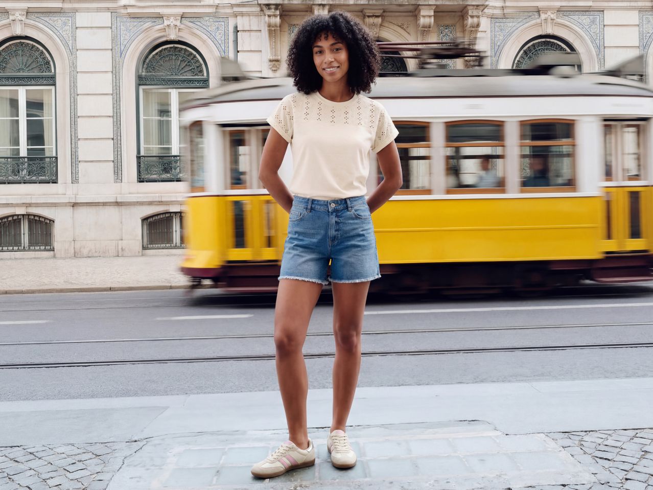 Woman in a cream top and denim shorts, standing on a street with a yellow tram passing by.