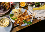 Grilled shrimp skewers, pita bread, hummus, BBQ chicken, and a beet salad on a wooden table.