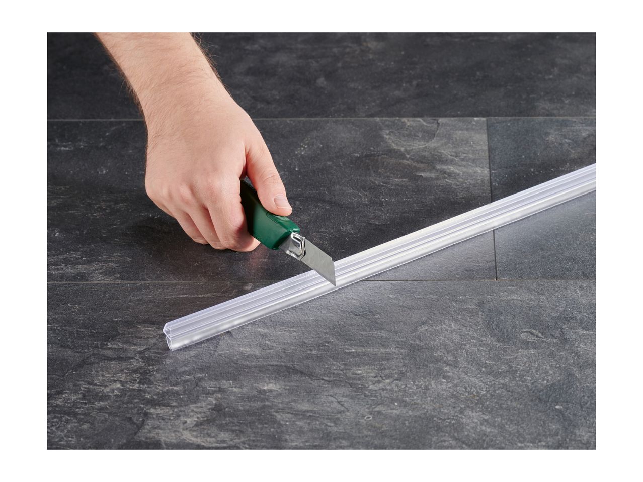Hand cutting a transparent plastic strip with a utility knife on a dark tiled surface.