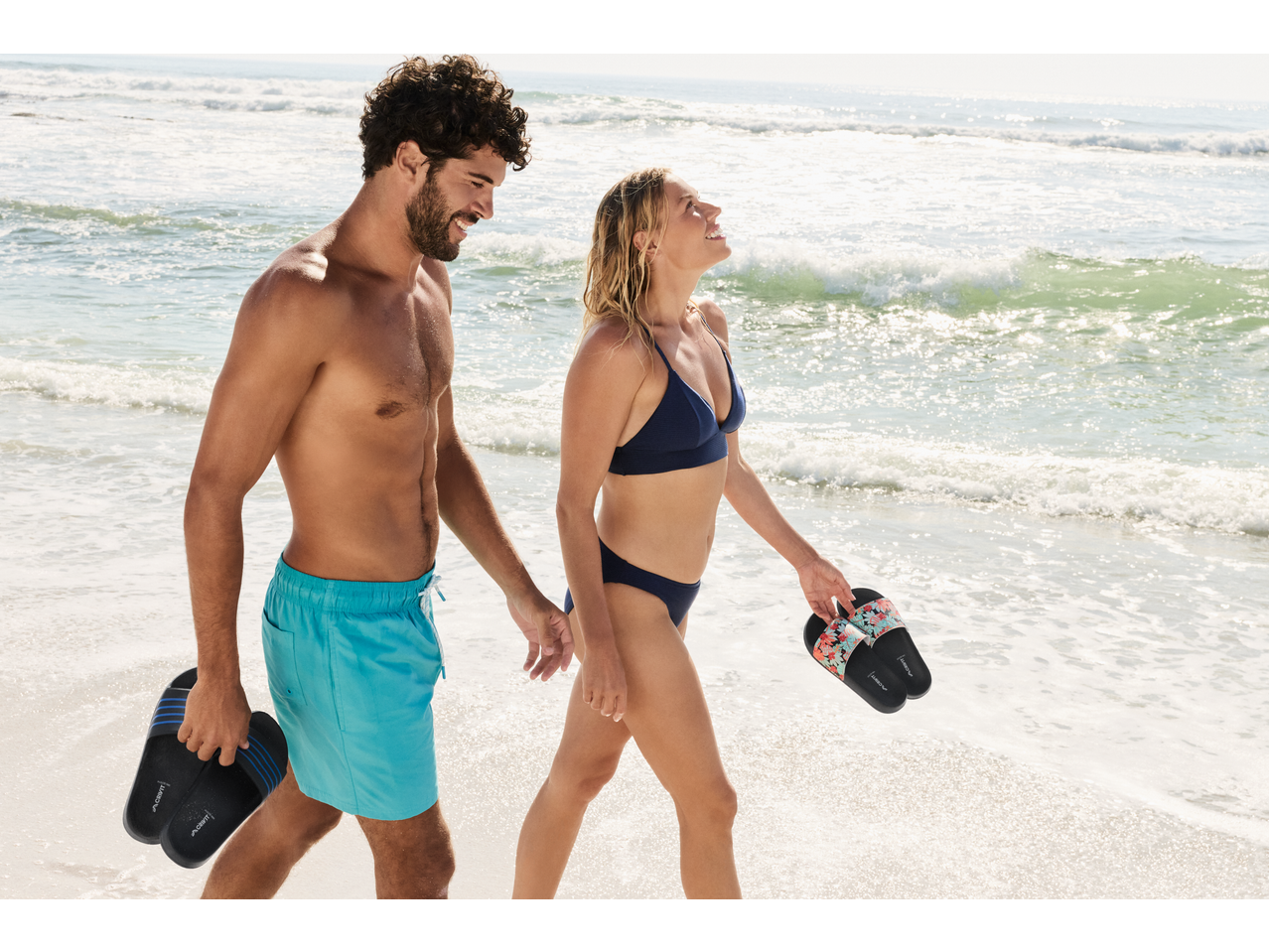 A couple walks on the beach, holding slides. The man wears blue swim shorts, and the woman wears a navy bikini.