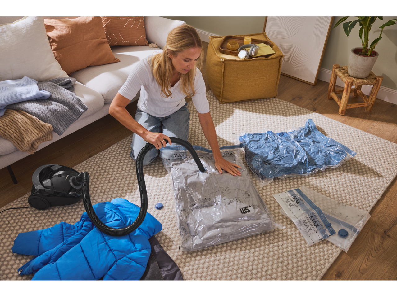 Woman using a vacuum cleaner to vacuum pack clothes into storage bags on a rug.