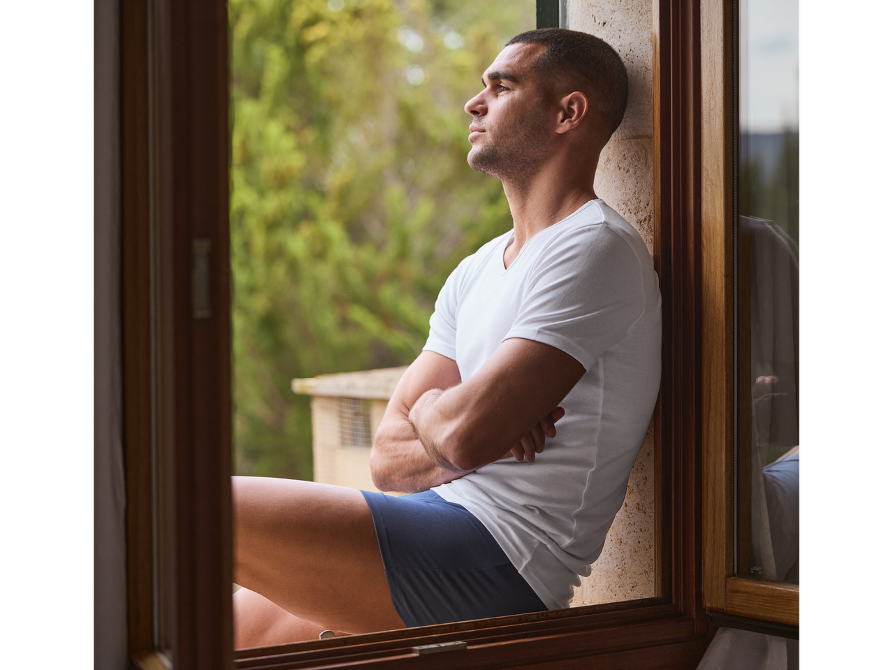 A man sits by a window wearing underwear and a t-shirt.