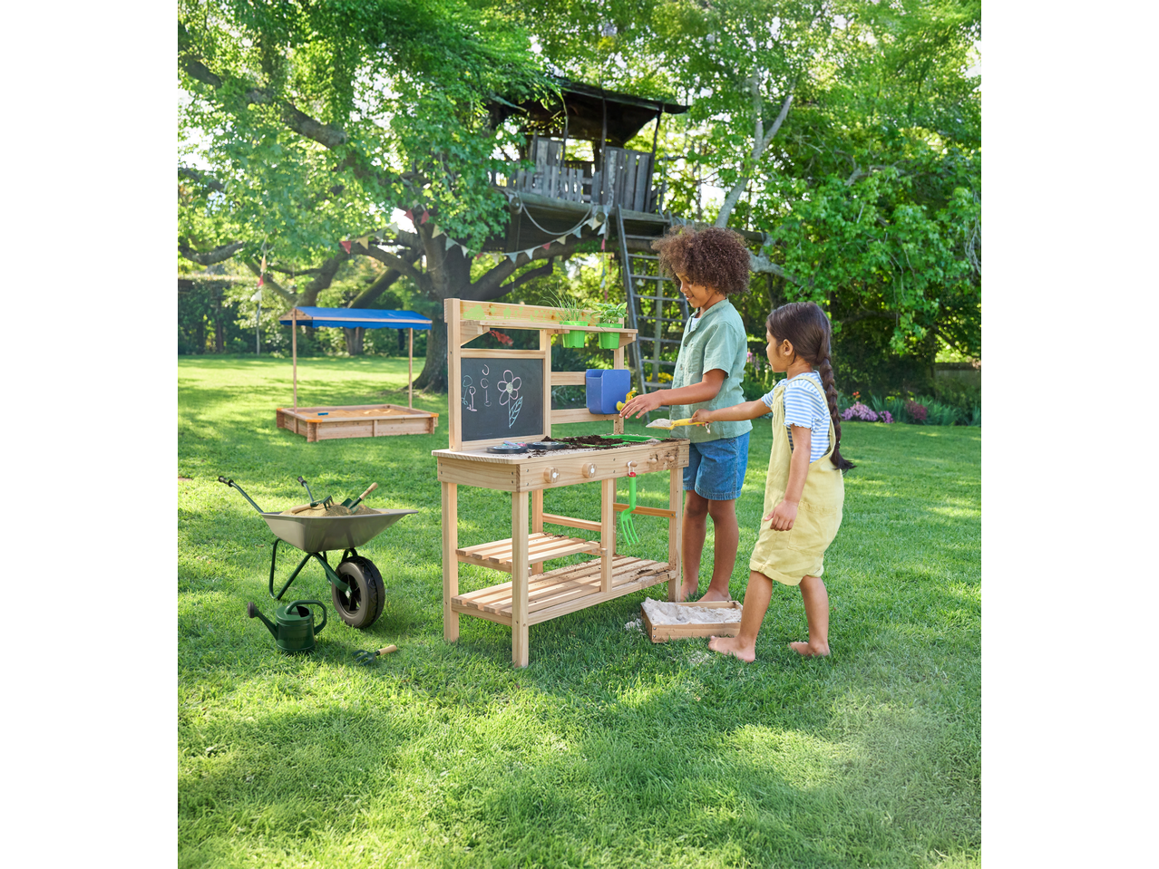 Crianças a brincar no jardim com mesa de jardinagem de madeira e carrinho de mão com ferramentas.