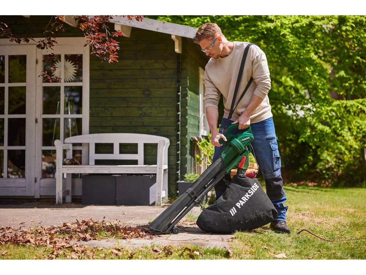 Man using a Parkside leaf blower to clear leaves from a patio next to a green shed.