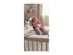 Baby in a pink outfit sitting in a grey baby bouncer in a nursery with shelves and toys.