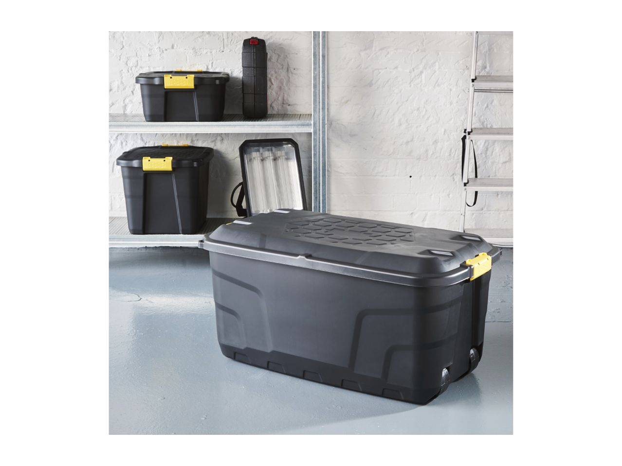 Black storage boxes and toolboxes on shelves and floor in a garage.