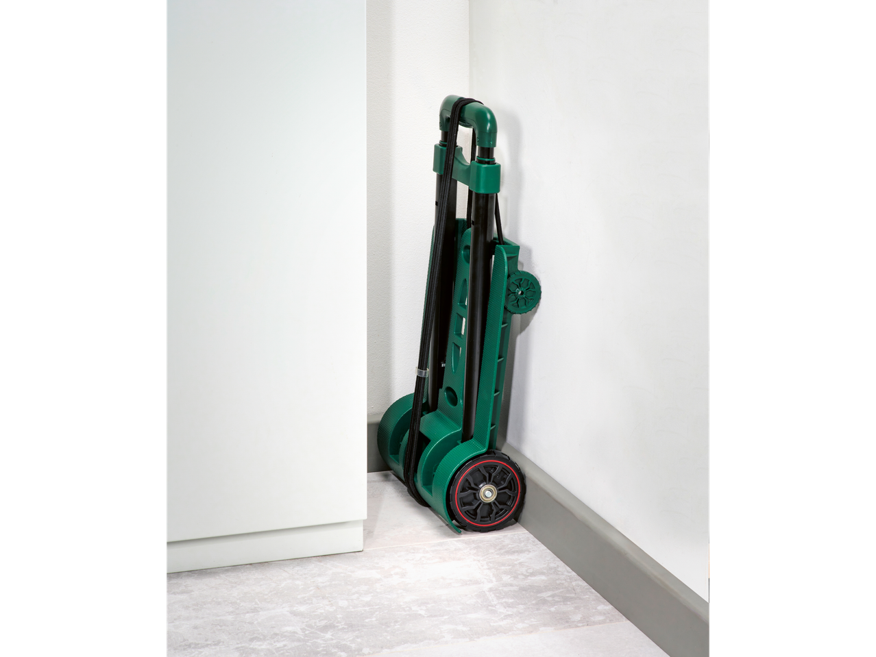 A green folding hand truck is stored upright in a corner against a white wall.