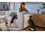 Mulher sorridente a observar um gato a arranhar um Zoofari® Kratzmatte num sofá.